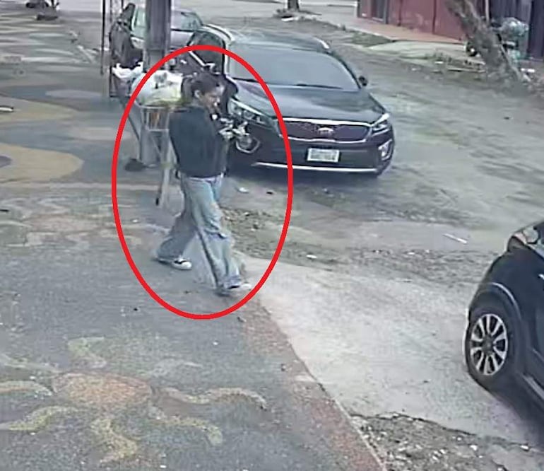 Joven con chaqueta negra y jeans claros camina sola por calle, sosteniendo un objeto, en un ambiente urbano vacío.
