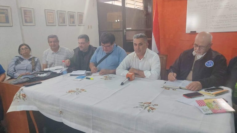 Silvio Piris en camisa blanca y Rafael Resquín en suéter azul, rodeados de cuatro personas en reunión formal con botella de agua en mesa.