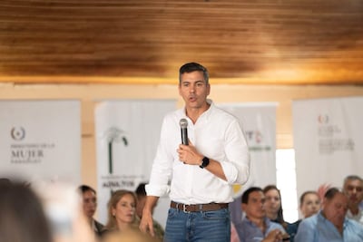 Santiago Peña en camisa blanca y jeans, sosteniendo un micrófono mientras habla a una audiencia, con Leticia Ocampo atendiendo.