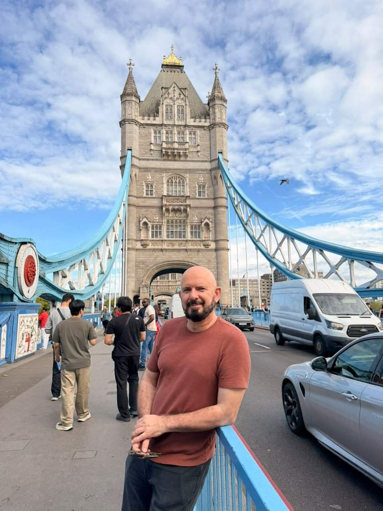 Hombre de cabello corto y barba, con camiseta terracota, posando junto a una barandilla azul frente al puente.