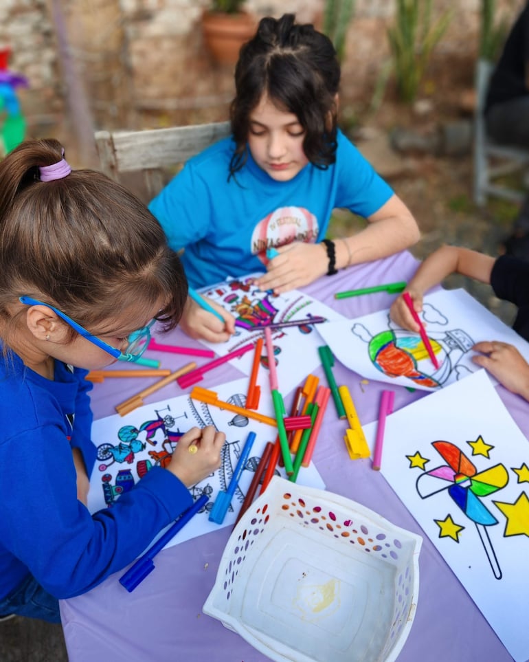 Niños con camisetas azules concentran en dibujos sobre una mesa, rodeados de marcadores y una cesta vacía.