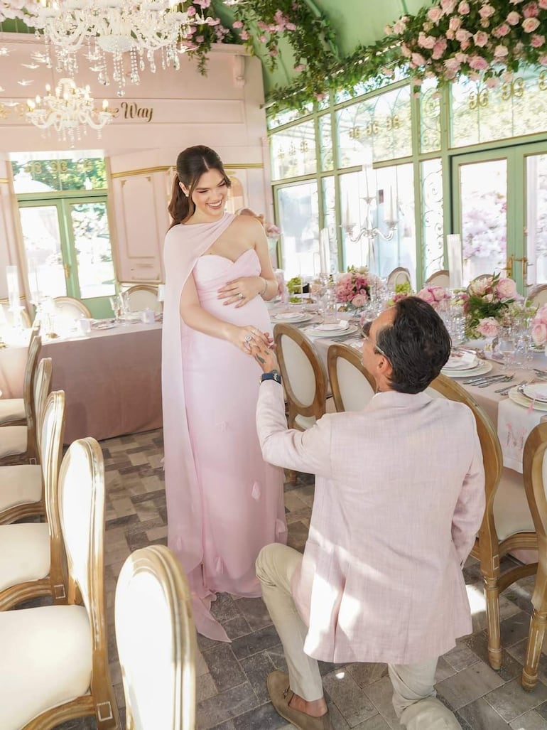 Nadia Ferreira en vestido rosa claro sonríe, mientras Marc Anthony arrodillado frente a ella, vestido con saco rosa claro, en ambiente decorado.