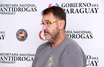 Hombre de barba y gafas con camiseta gris, de perfil frente a un panel de la Secretaría Nacional Antidrogas del Paraguay.