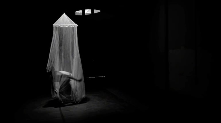 Artista en escena oscura, movimiento sutil con tela blanca, creando atmósfera performática y reflexiva sobre la maternidad forzada.