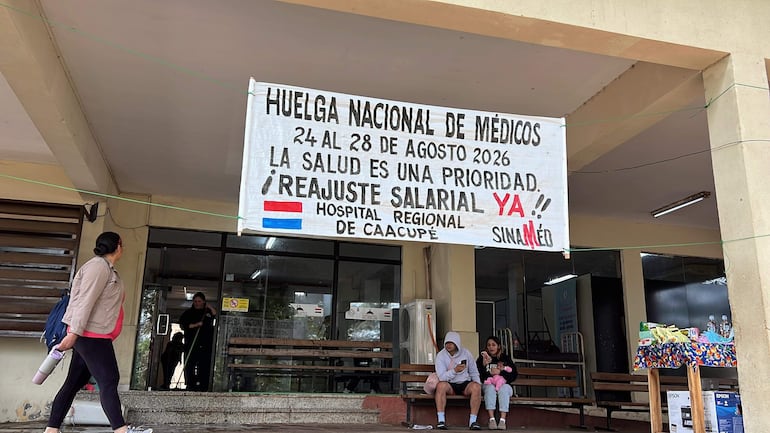 Mujer de espaldas y hombre sentado con teléfono, pancarta en el hospital pidiendo reajuste salarial, ambiente de protesta pacífica.