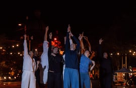 Mujer en ropa blanca levanta el puño mientras otros a su lado hacen lo mismo en un ambiente festivo iluminado suavemente por luces colgantes.