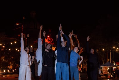 Mujer en ropa blanca levanta el puño mientras otros a su lado hacen lo mismo en un ambiente festivo iluminado suavemente por luces colgantes.