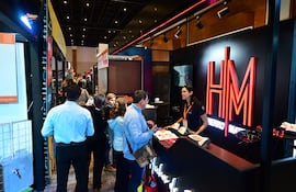 Mujer de pie en un mostrador, hablando con un hombre de chaqueta clara en un ambiente iluminado de feria comercial.
