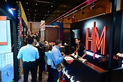 Mujer de pie en un mostrador, hablando con un hombre de chaqueta clara en un ambiente iluminado de feria comercial.