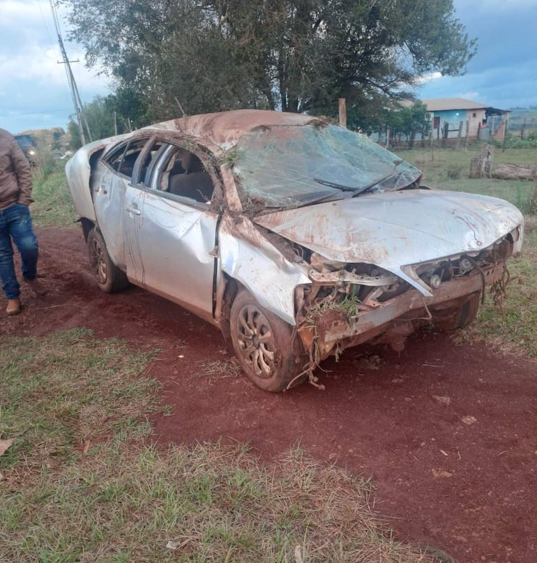 Hombre con chaqueta marrón y jeans camina cerca de un coche plateado destrozado en un paisaje rural.