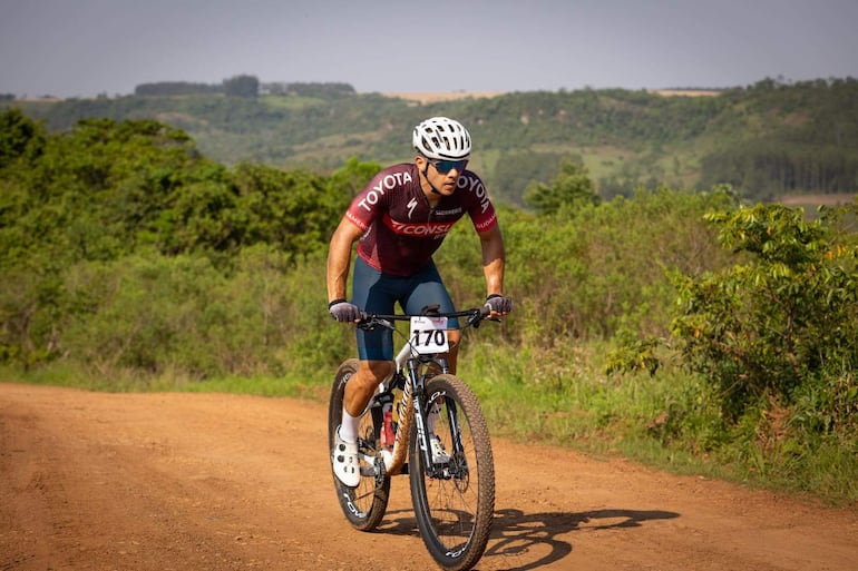 Ciclista con casco blanco y camisete rojo y azul, avanza en carretera de tierra, rodeado de colinas verdes.