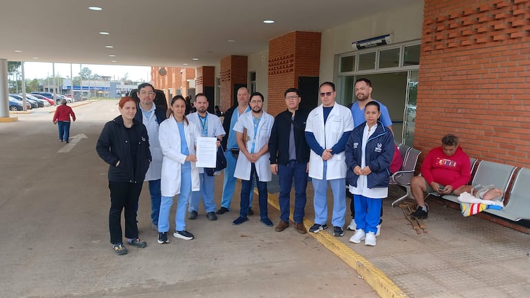 Grupo de médicos en batas blancas y uniformes azules se agrupan frente a un hospital durante una protesta, uno sostiene un papel.