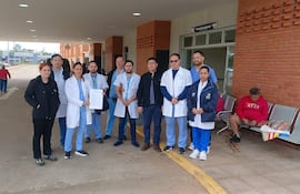 Grupo de médicos en batas blancas y uniformes azules se agrupan frente a un hospital durante una protesta, uno sostiene un papel.