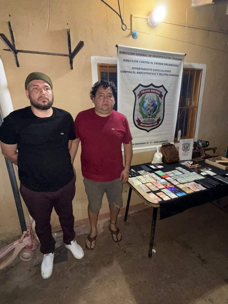 Dos hombres detenidos en el recinto de la Policía Nacional, uno con camiseta negra y otro con camiseta roja, rodeados de documentos.