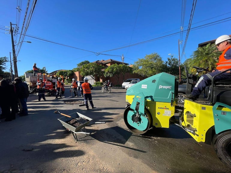 Grupo de trabajadores con chalecos naranjas y cascos operando maquinaria en la calle, rodeados de árboles y un camión.