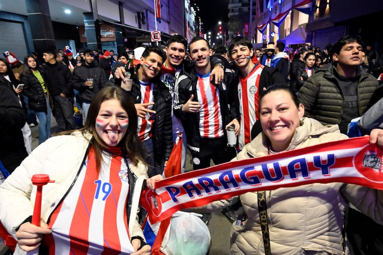 Joven sonriente con rostro pintado en colores de Paraguay y camiseta, junto a dos hombres con banderas en un ambiente festivo.