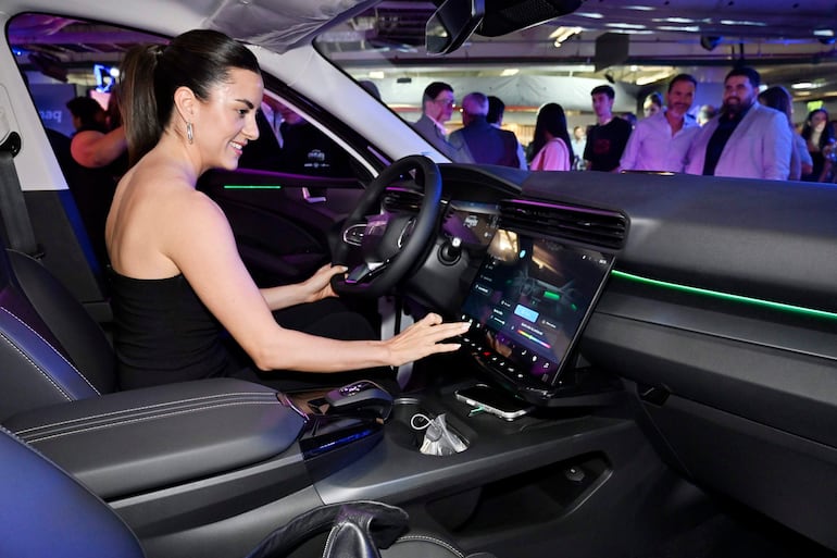 Mujer con blusa negra y cabello recogido al volante de un vehículo, concentrada en la pantalla táctil del tablero. Personas observan detrás.