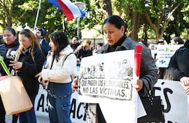 Mujer con cartel que dice 'QUE PAGUEN LOS RESPONSABLES, NO LAS VÍCTIMAS', rodeada de manifestantes y vegetación en un parque.