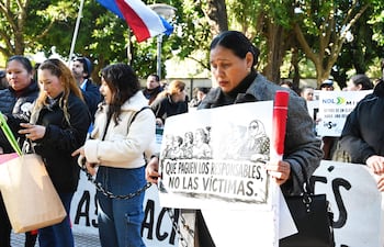 Mujer con cartel que dice 'QUE PAGUEN LOS RESPONSABLES, NO LAS VÍCTIMAS', rodeada de manifestantes y vegetación en un parque.
