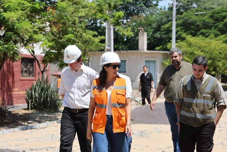 Mujer con chaleco naranja y casco de seguridad habla con cuatro hombres en sitio de construcción rodeado de vegetación bajo sol.