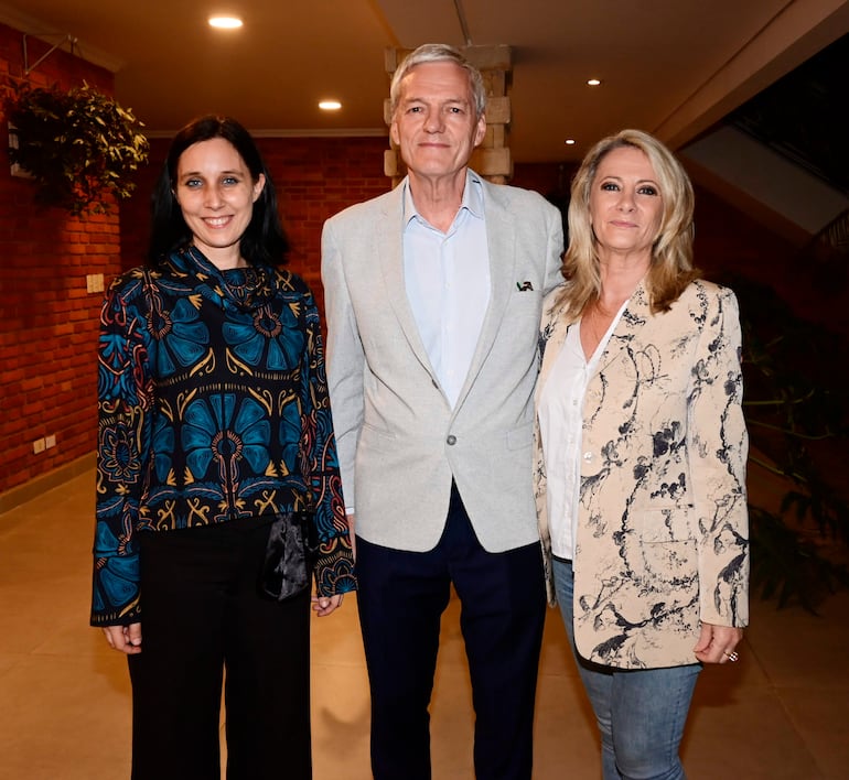 Mujer de cabello largo con blusa colorida, hombre canoso en chaqueta clara y mujer rubia con chaqueta clara, todos sonriendo en ambiente acogedor.