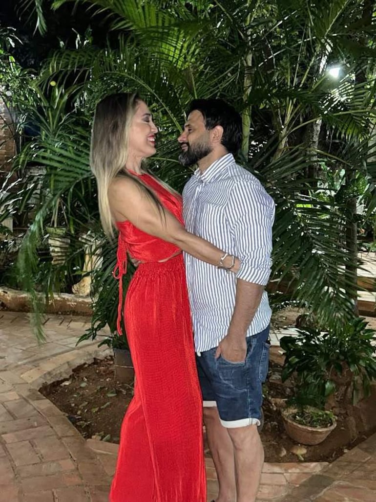 Mujer con vestido rojo y hombre con camisa de rayas, sonriendo juntos en un jardín con vegetación densa.
