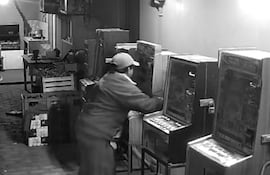 Hombre de 42 años, vestido de trabajo y gorra, manipulando una máquina tragamonedas en un salón desordenado.