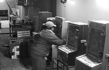 Hombre de 42 años, vestido de trabajo y gorra, manipulando una máquina tragamonedas en un salón desordenado.