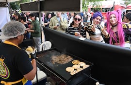 Hombre con gorra y delantal amarillo cocina carne en parrilla; tres mujeres sonríen, una con pelo púrpura y otra con gafas de sol.