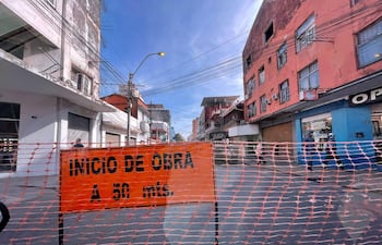 Señal de 'INICIO DE OBRA' en cruce urbano, con malla naranja que limita el acceso. Calle despejada y edificios de tonos grises y rojos.
