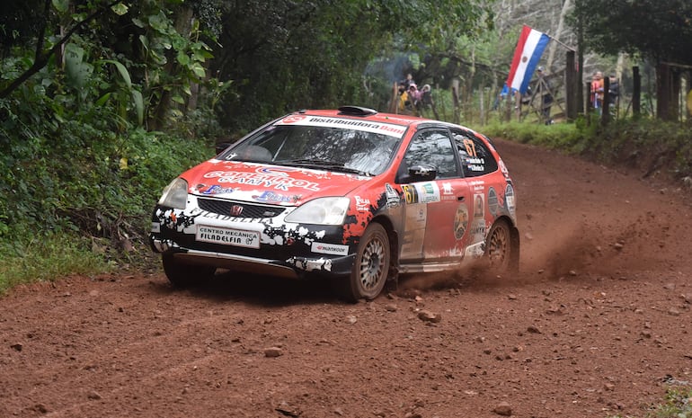 Automóvil Honda de rally en movimiento, cubierto de barro, rodeado de vegetación y con una bandera paraguaya al fondo.
