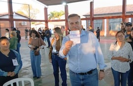 Hombre canoso sostiene documento al centro, rodeado de votantes en ambiente bien iluminado y participativo en Pilar.
