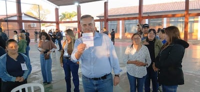 Hombre canoso sostiene documento al centro, rodeado de votantes en ambiente bien iluminado y participativo en Pilar.