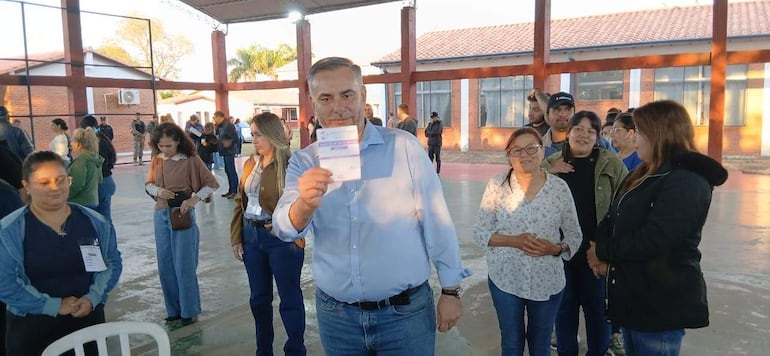 Hombre canoso sostiene documento al centro, rodeado de votantes en ambiente bien iluminado y participativo en Pilar.