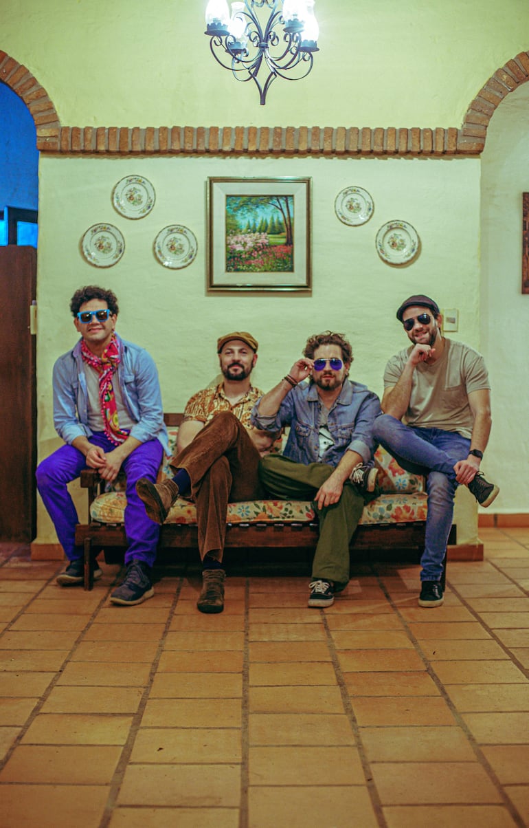 Cuatro hombres conversan en un sofá; uno lleva camisa de denim y pantalones morados, otro vestimenta colorida y la atmósfera es acogedora.