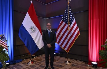 Hombre en traje oscuro y corbata azul sonríe frente a las banderas de Paraguay y Estados Unidos en un evento formal.