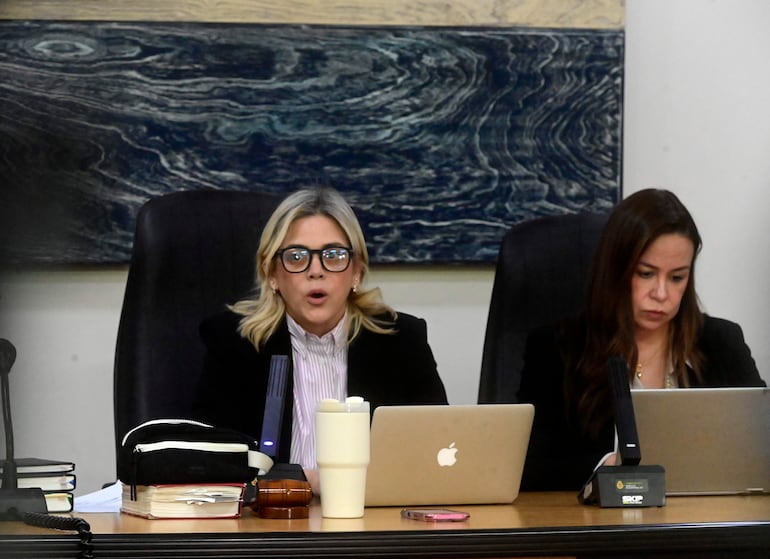 Mujer rubia en camisa blanca con rayas habla mientras una mujer castaña, con laptop, parece concentrada en la discusión.
