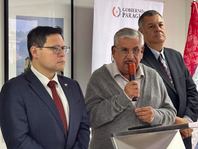Tres hombres en trajes formales, el central con micrófono, banderas de Paraguay y cartel del Gobierno de Paraguay en el fondo.