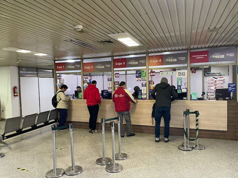 Cuatro personas en fila en mostrador, una lleva sudadera roja y otra abrigo claro, en ambiente de oficina pública.