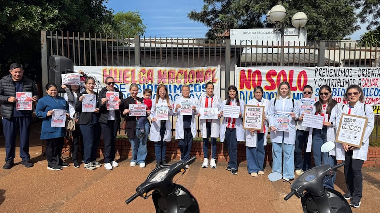 Grupo de bioquímicos en jalecos blancos sostiene carteles durante protesta, una mujer con niño destaca en la primera fila.