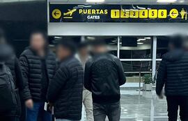 Seis migrantes en el aeropuerto, vestidos con chaquetas oscuras, esperando en un área de embarque con cartel visible de 'PUERTAS'.