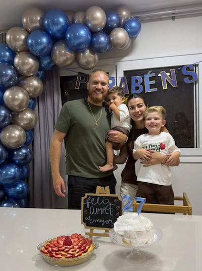 Isidro Pitta sonriente sostiene a un niño mientras una mujer lo acompaña junto a otro niño, decoraciones de cumpleaños al fondo.