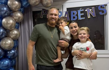 Isidro Pitta sonriente sostiene a un niño mientras una mujer lo acompaña junto a otro niño, decoraciones de cumpleaños al fondo.