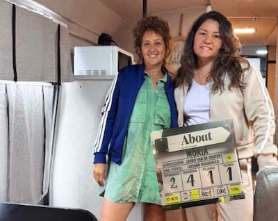 Shirley Giménez con chaqueta azul y camiseta verde, junto a Coral Gabaglio en chaqueta beige, sosteniendo cartel de producción.