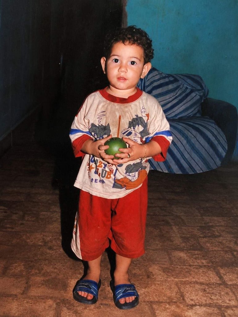 Niño pequeño con camiseta clara y pantalones cortos rojos, sosteniendo una fruta verde en un ambiente acogedor.