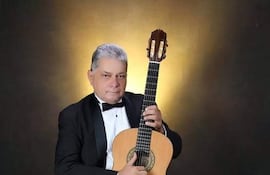 Gabriel Tapia sentado en traje negro con pajarita blanca, sosteniendo guitarra acústica, iluminado con luz suave en fondo simple.