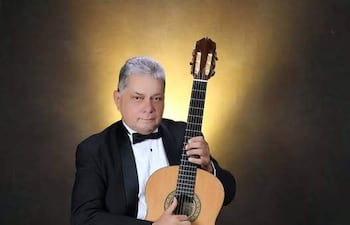 Gabriel Tapia sentado en traje negro con pajarita blanca, sosteniendo guitarra acústica, iluminado con luz suave en fondo simple.