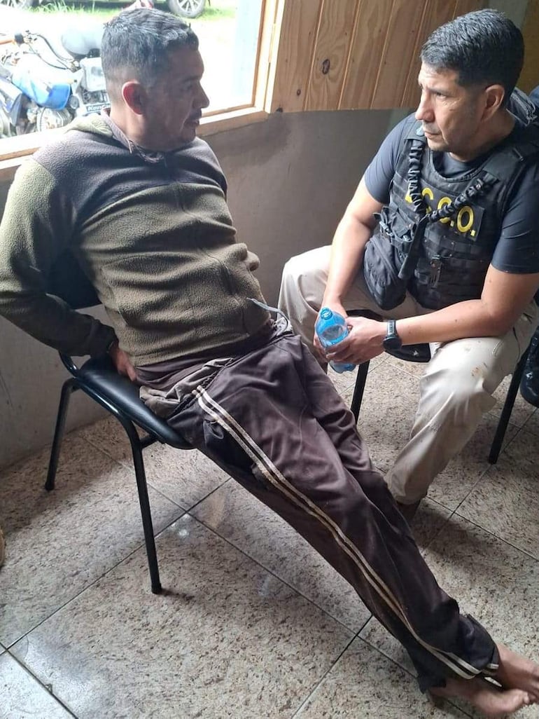 Dos hombres sentados en habitación: uno escucha atentamente mientras el otro sostiene una botella de agua, ambos interactuando.