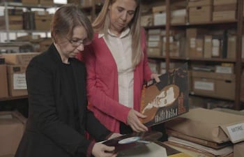 Mujer de cabello corto y gris observa un disco, mientras otra con cabello rubio revisa documentos en una biblioteca llena de cajas.