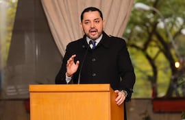Luis Bello gesticula en podio con abrigo negro y corbata morada, frente a un ambiente formal con vegetación de fondo.